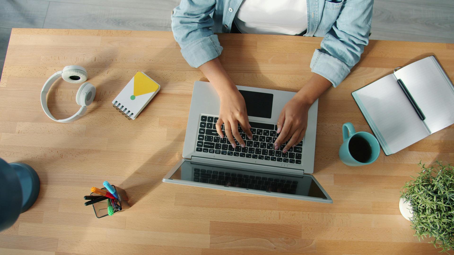 An overhead view of a desk — hands at a laptop, with an open notebook beside it, its pages still blank
