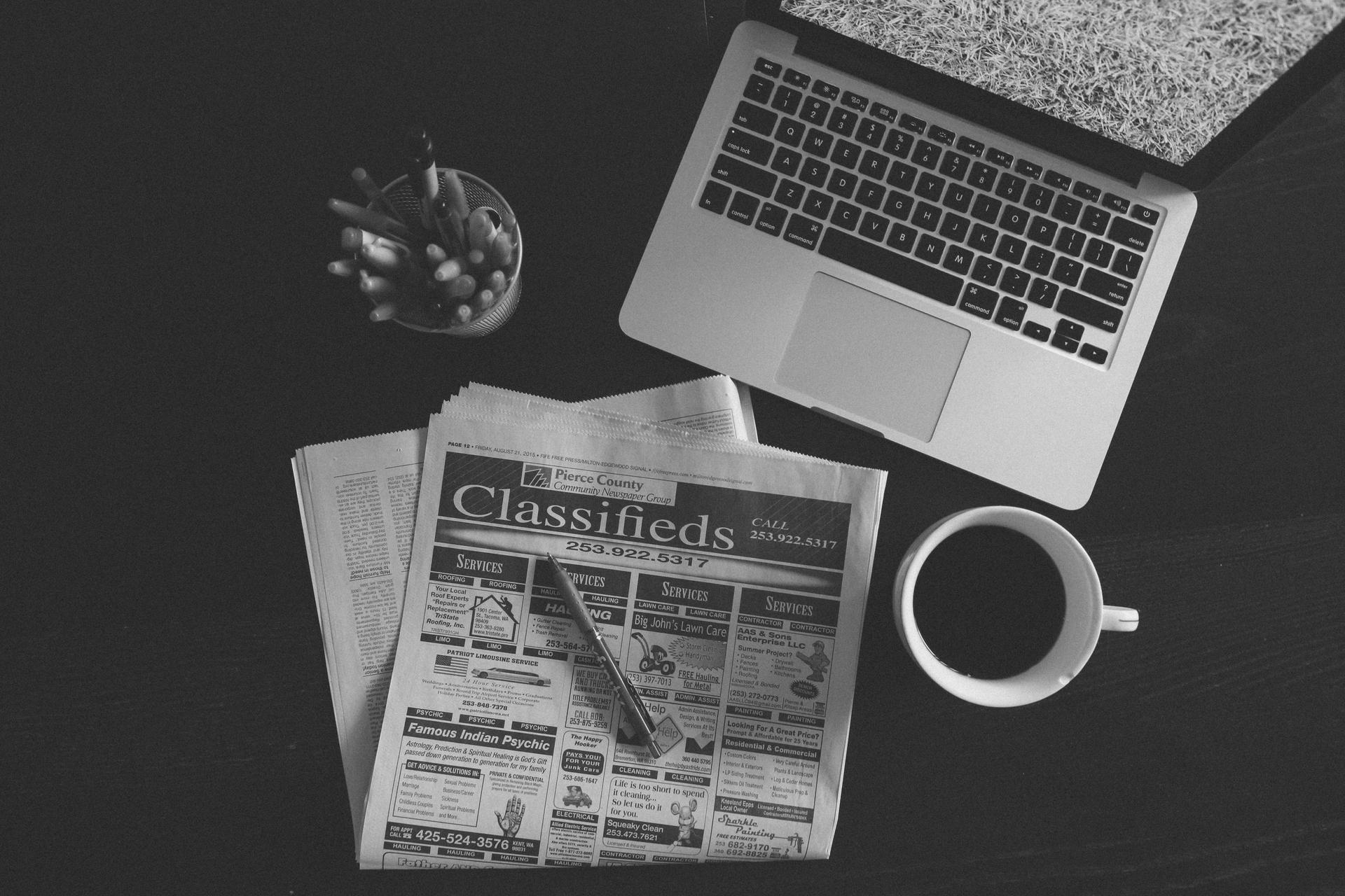 A black-and-white overhead shot of a folded newspaper, an open laptop, a jar of pens and a cup of black coffee on a dark desk